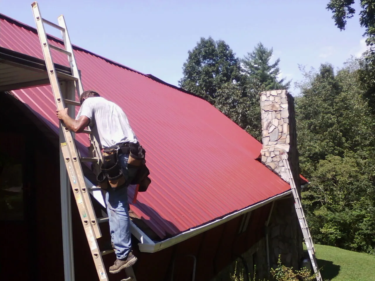 Roofer on a ladder beside a red metal roof and stone chimney — Soffit & Fascia Repair by Roofing Brothers in Fishs Eddy
